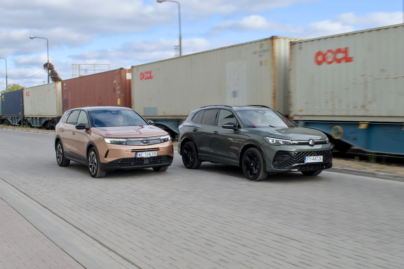 Dwa nowoczesne samochody SUV jadą obok siebie po miejskiej ulicy, w tle widoczne kontenery transportowe na wagonach kolejowych i niebo z lekkim zachmurzeniem.