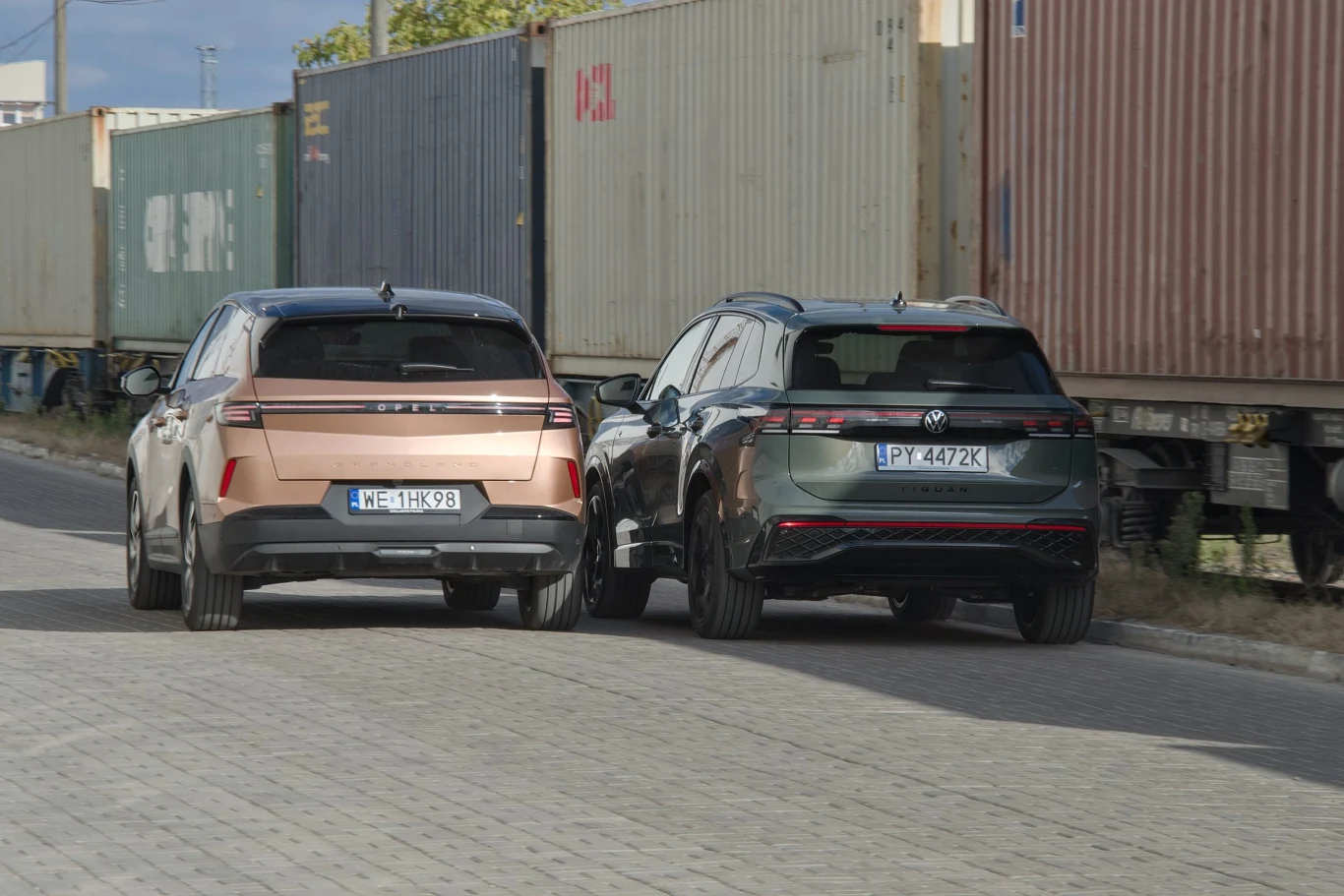 Dwa nowoczesne SUV-y zaparkowane obok siebie na brukowanej ulicy, w tle rząd kontenerów transportowych na wagonach kolejowych.
