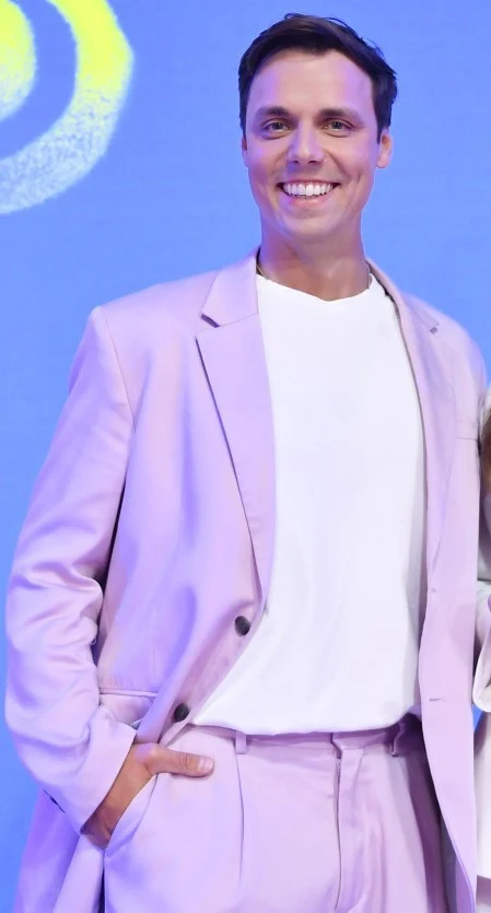 Mateusz Jędraś Mężczyzna uśmiechnięty, ubrany w jasnoróżowy garnitur i białą koszulkę, stoi na tle niebieskiej ściany z żółtym wzorem w rogu.