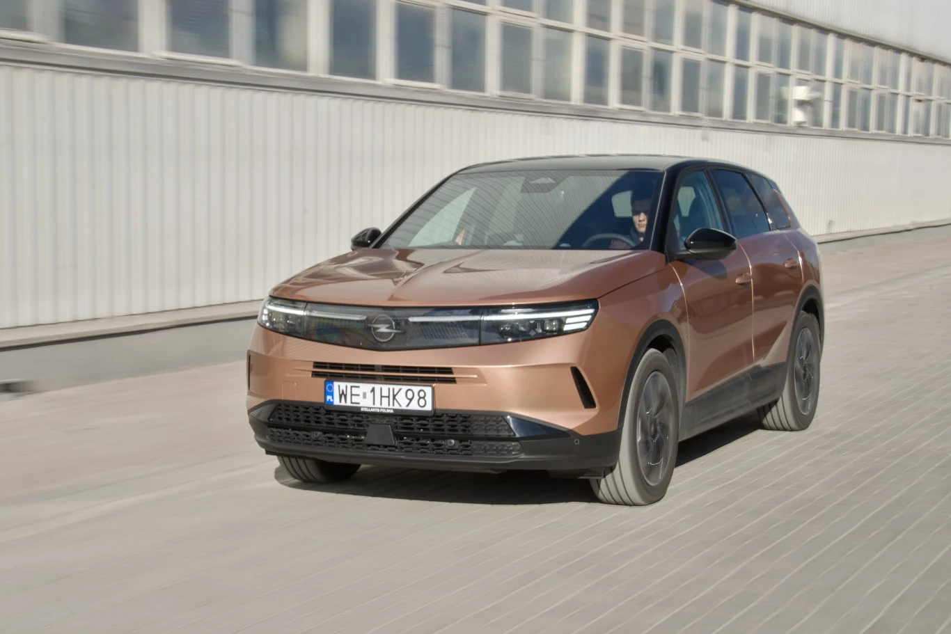 Nowoczesny SUV jadący ulicą w jasnej okolicy przemysłowej, auto w kolorze miedzianym z polskimi tablicami rejestracyjnymi, dynamiczne ujęcie podkreślające ruch pojazdu.