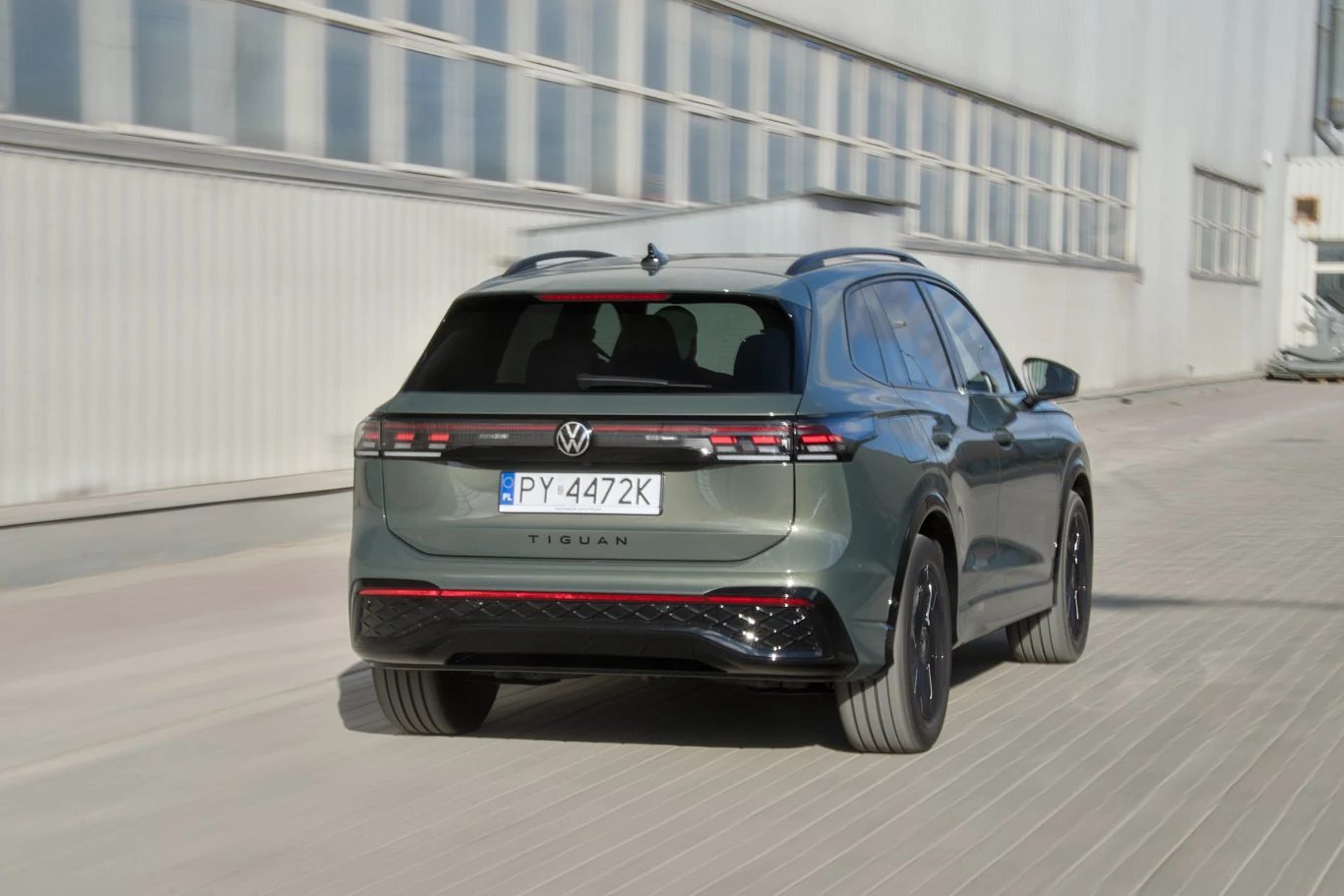 Szary SUV Volkswagen Tiguan widoczny od tyłu porusza się wzdłuż betonowej drogi przy jasnym, przemysłowym budynku. Na samochodzie czytelny jest numer rejestracyjny oraz logo producenta.