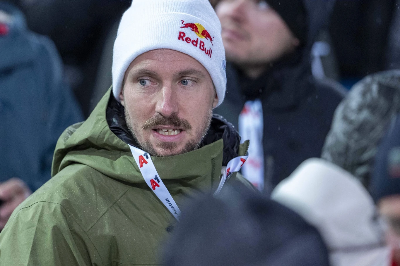 Marcel Hirscher Mężczyzna w białej czapce z logo Red Bull oraz kurtce w kolorze oliwkowym, z identyfikatorem zawieszonym na szyi, znajduje się w tłumie ludzi ubranych w zimowe ubrania.