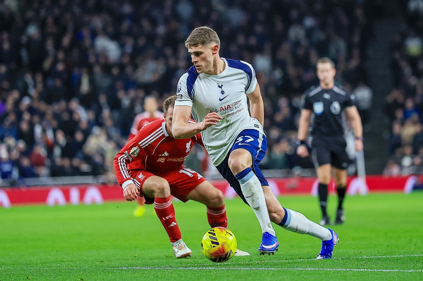 Micky van de Ven Dwóch piłkarzy rywalizuje o piłkę na zielonej murawie podczas meczu, wokół widać publiczność na trybunach i sędziego obserwującego grę.