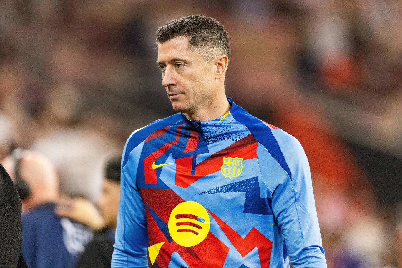 Robert Lewandowski Piłkarz w barwach FC Barcelony noszący kolorową bluzę treningową z logiem klubu oraz sponsora, skoncentrowany, znajduje się na tle nieostrego stadionu.