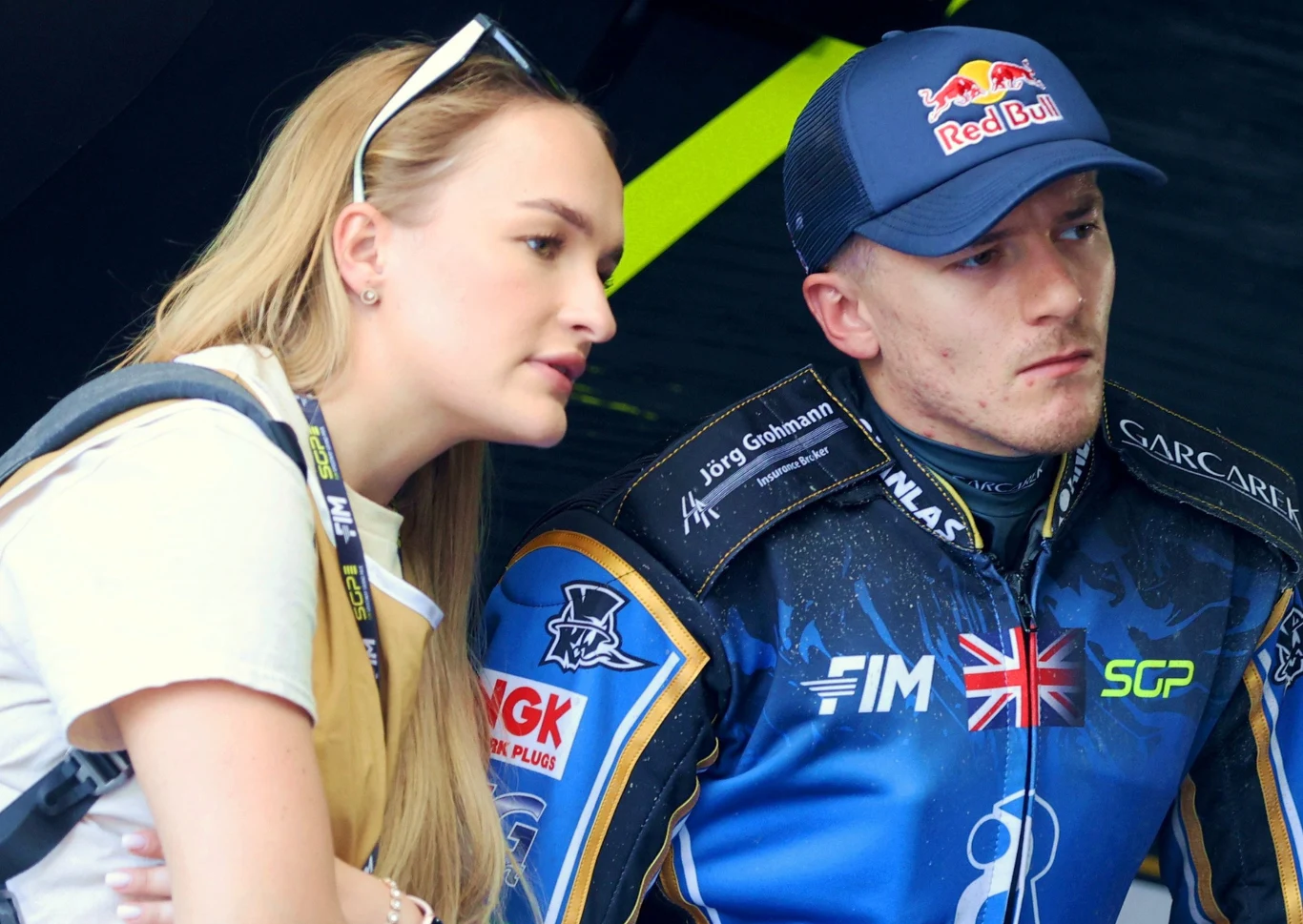 Kobieta w blond włosach pochyla się i rozmawia z motocyklistą ubranym w kombinezon sportowy oraz czapkę z logo Red Bull. Na twarzy motocyklisty widać skupienie i powagę.