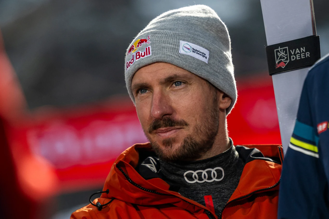 Marcel Hirscher Mężczyzna w szarej czapce z logo Red Bull, ubrany w czerwoną kurtkę sportową i czarną bluzę z logo Audi, patrzy w bok na tle rozmytego czerwonego banera. Twarz dobrze oświetlona, wyraz skupienia.