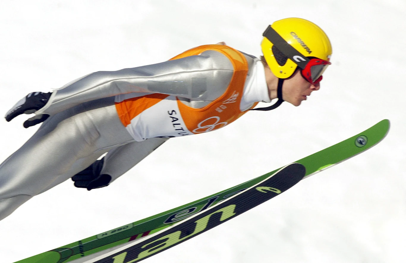 Zawodnik w kombinezonie narciarskim z kaskiem i goglami podczas skoku narciarskiego na tle śniegu, widoczny w locie na nartach.