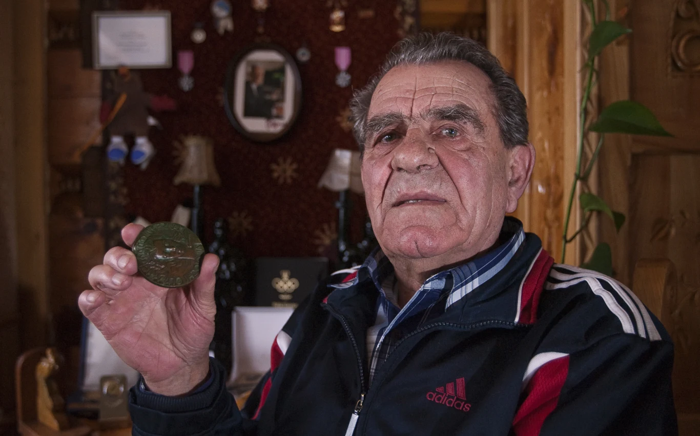 Starszy mężczyzna w sportowej kurtce trzyma w dłoni medal, w tle ozdobne i pamiątkowe przedmioty zawieszone na ścianie drewnianego wnętrza.