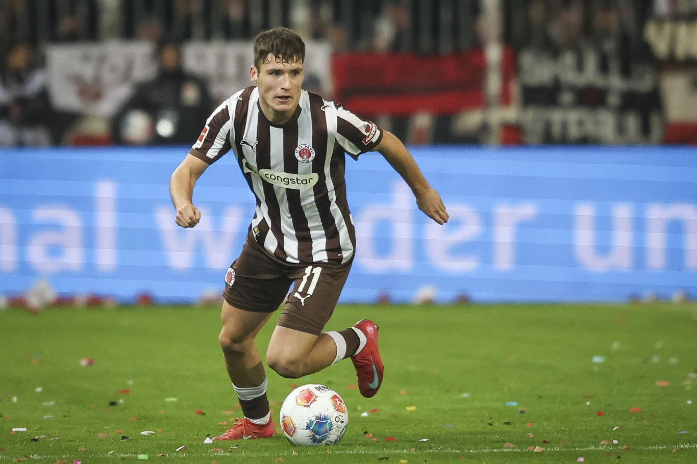 Piłkarz w pasiastym stroju sportowym biegnie z piłką po boisku, wokół widoczne są konfetti oraz rozmazane postacie kibiców na trybunach.