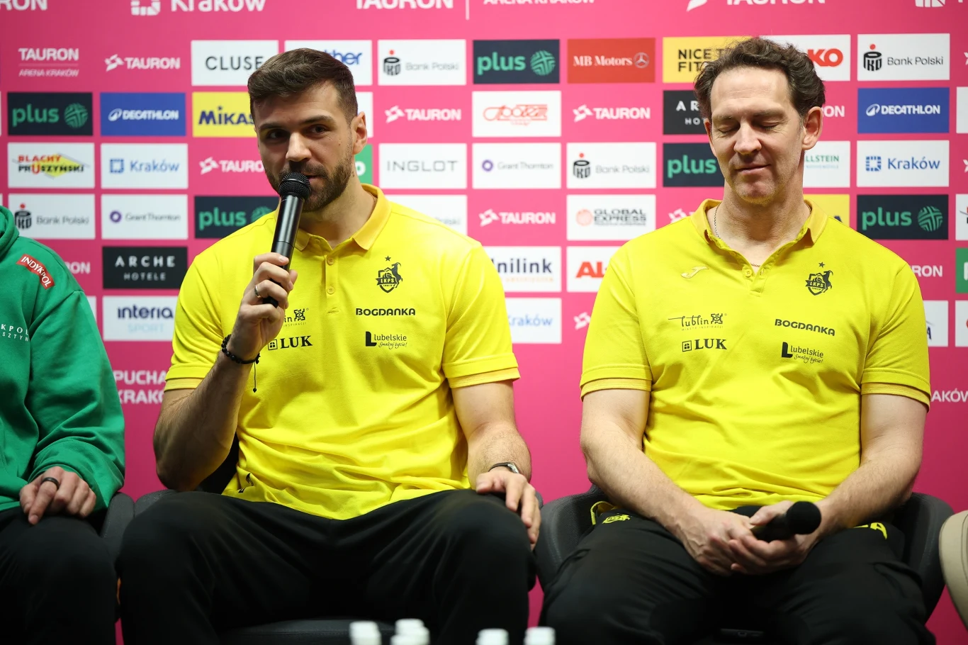 Dwóch mężczyzn w żółtych koszulkach klubowych siedzi obok siebie na tle ściany z logotypami sponsorów, jeden z nich trzyma mikrofon i przemawia, drugi trzyma ręce na kolanach.