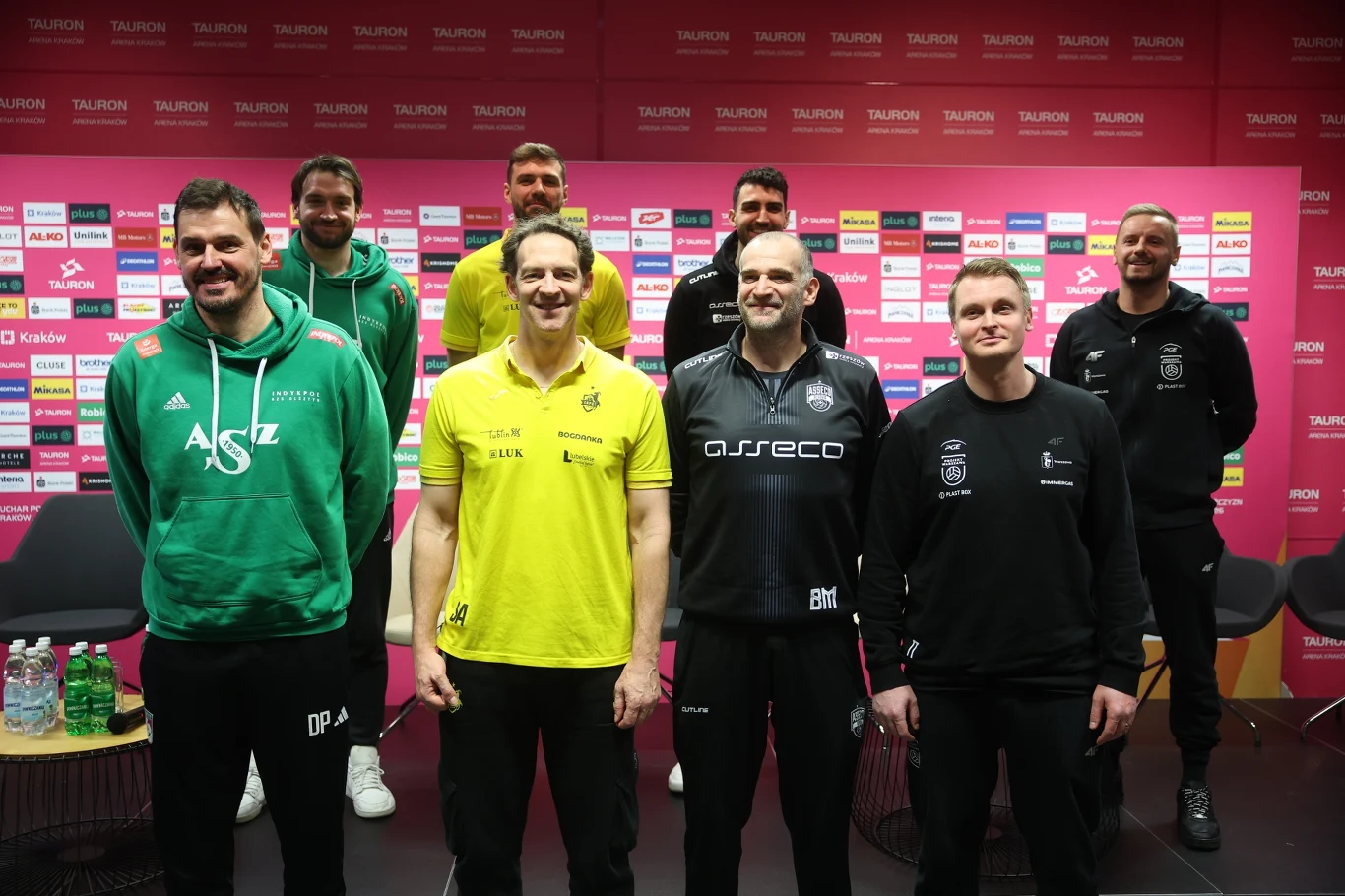 Grupa ośmiu mężczyzn ubranych w sportowe bluzy i koszulki z logo drużyn siatkarskich na tle ścianki sponsorskiej w sali konferencyjnej, na przodzie trzy osoby w jasnych i ciemnych sportowych strojach.
