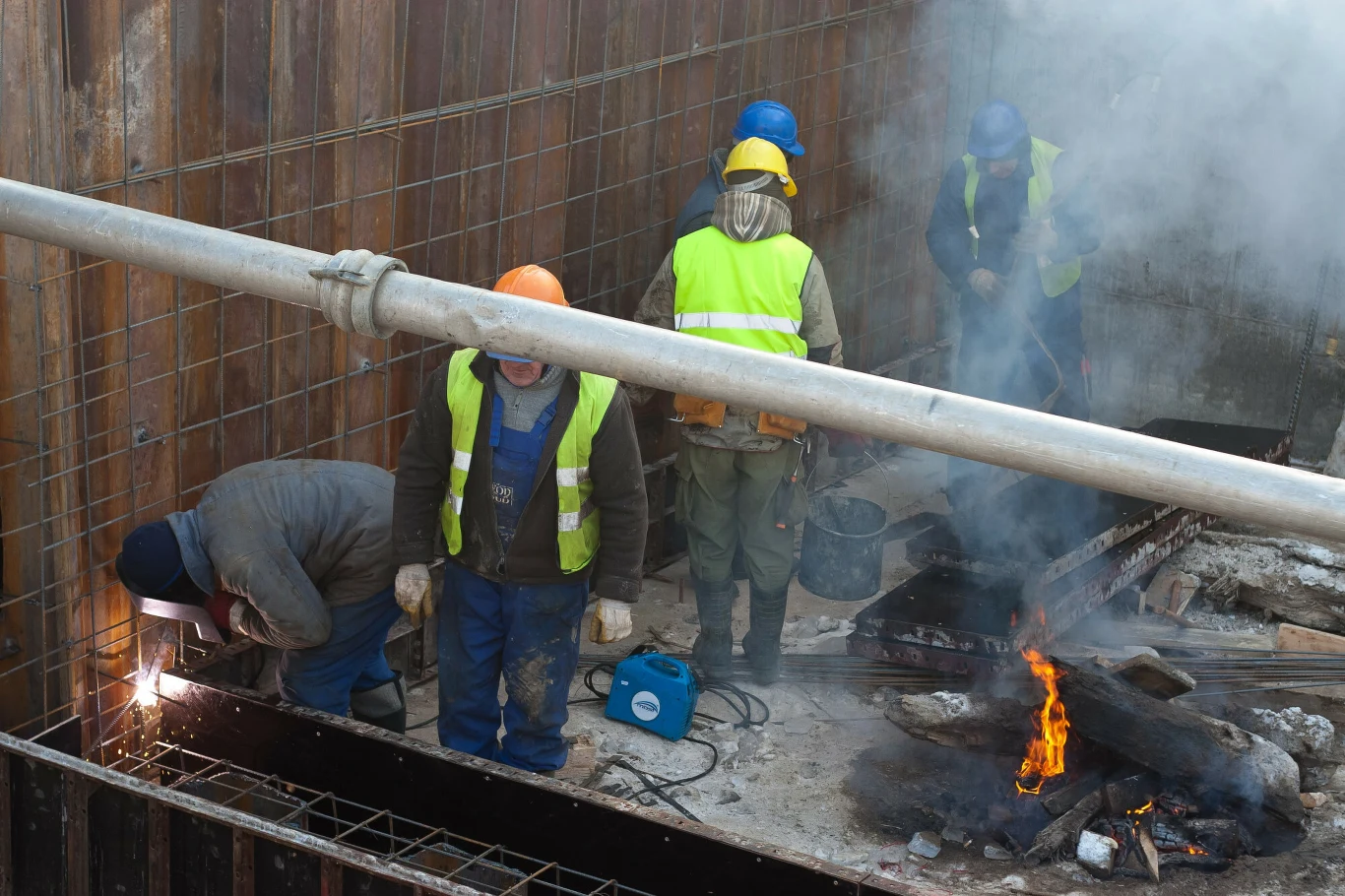 PIP jednak będzie mogła przekształcać umowy cywilnoprawne w etaty. Czterech robotników w odzieży ochronnej i kaskach pracuje na terenie budowy. Jeden z nich wykonuje prace spawalnicze, a obok pali się ognisko. W tle widoczne są elementy konstrukcji żelbetowych i metalowa siatka.