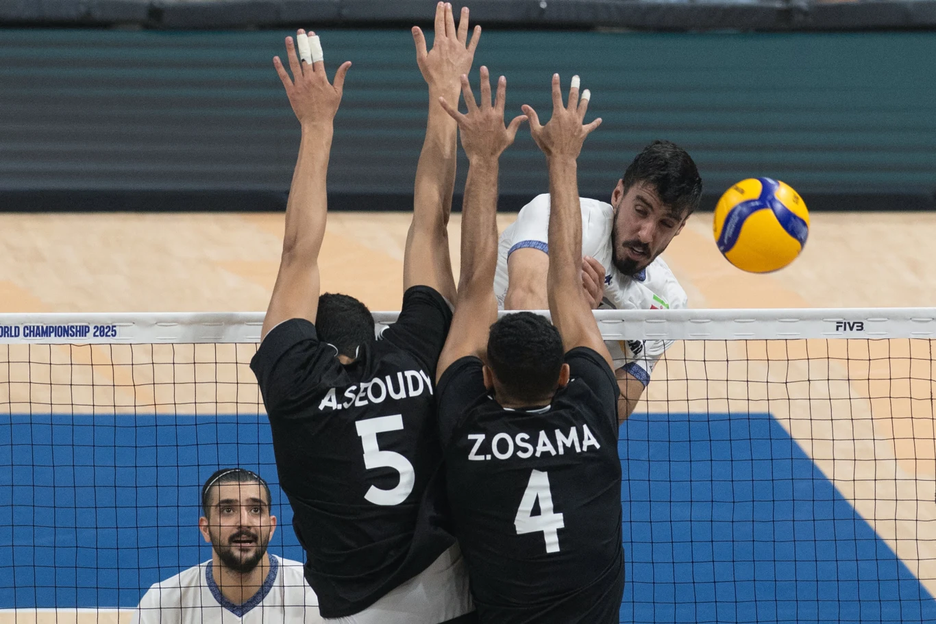 Zawodnik atakuje piłkę siatkową nad siatką, podczas gdy dwóch przeciwników w czarnych koszulkach próbuje ją zablokować wyciągając ręce ponad siatkę, w tle widoczny jeszcze jeden gracz.