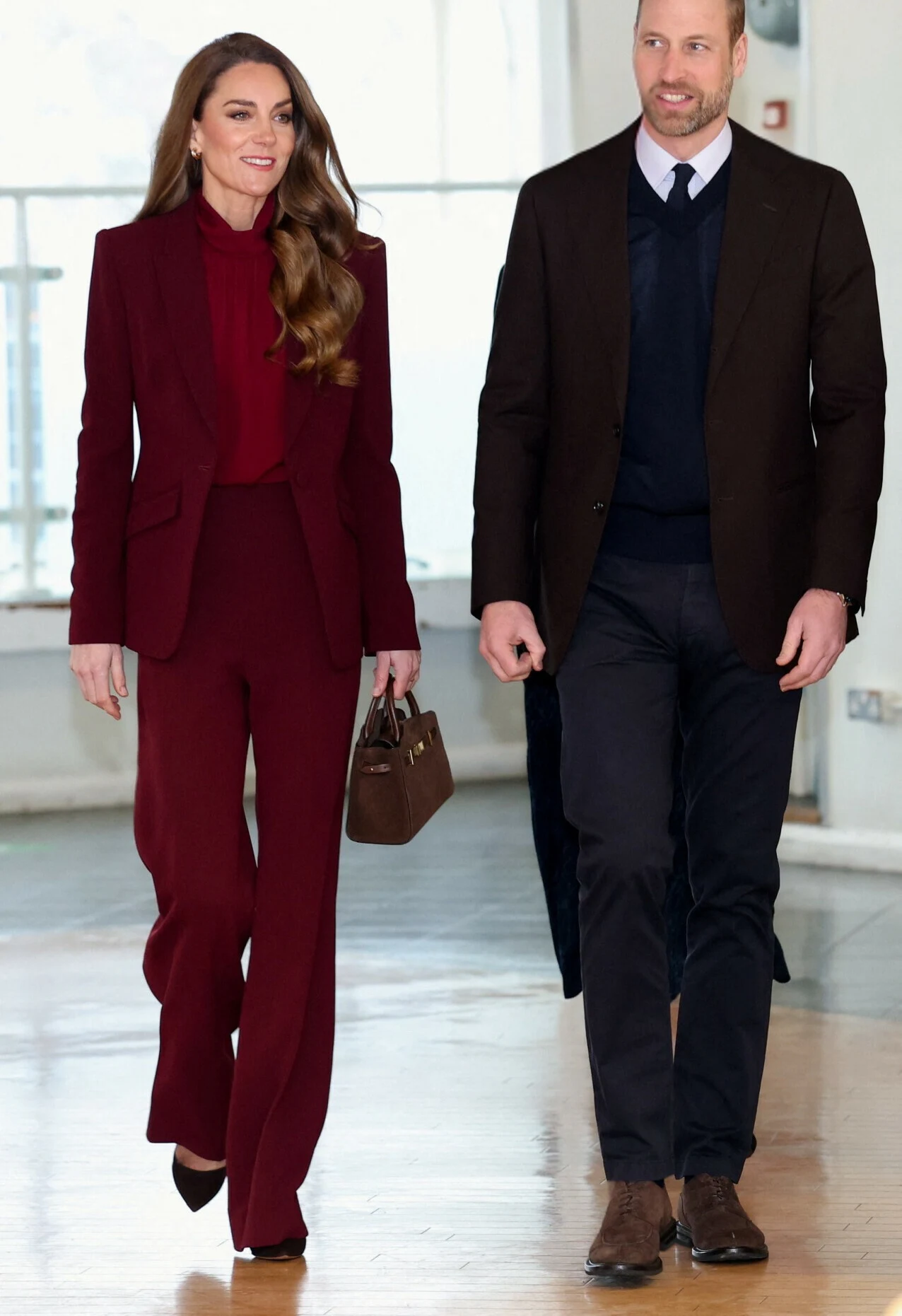 Elegancka kobieta w bordowym garniturze idzie obok mężczyzny ubranego półformalnie w ciemne spodnie, granatowy sweter i brązową marynarkę, oboje uśmiechnięci, kobieta trzyma brązową torebkę.
