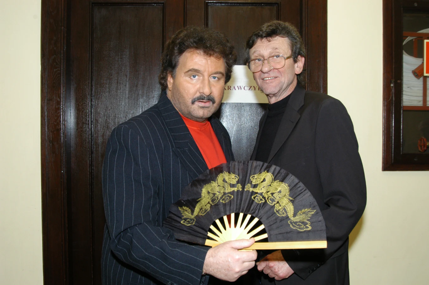 Dwóch mężczyzn pozuje w eleganckich garniturach na tle ciemnych drzwi, jeden z nich trzyma w ręce ozdobny wachlarz z motywem smoków.