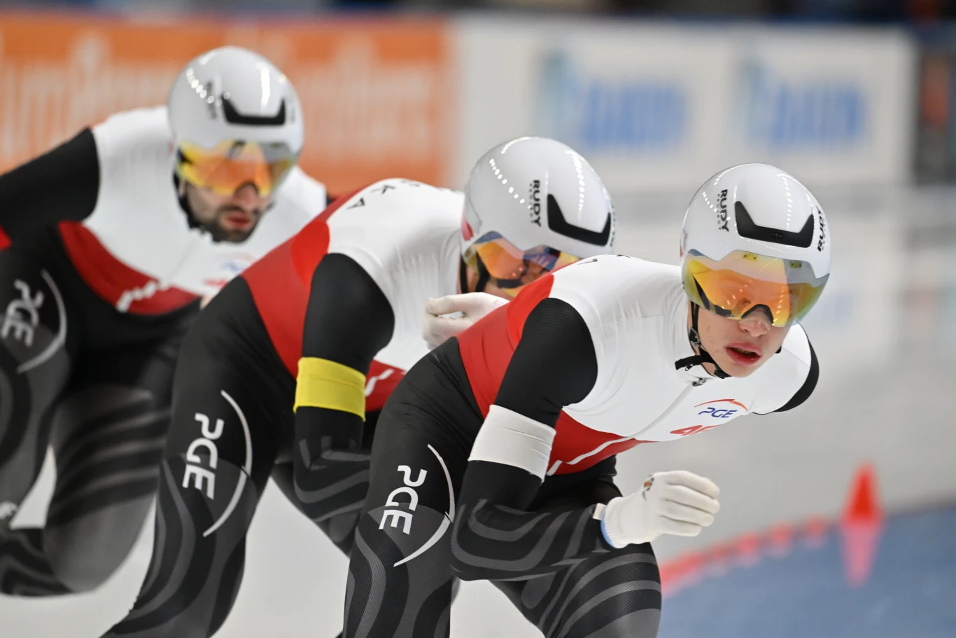 Władimir Siemirunnij, Szymon Palka i Marcin Bachanek Trzech panczenistów w profesjonalnych kombinezonach i kaskach jedzie po lodowej tafli toru, wszyscy pochylają się w dynamicznej pozycji, skupieni na zawodach, na ramionach widoczne logo sponsora.