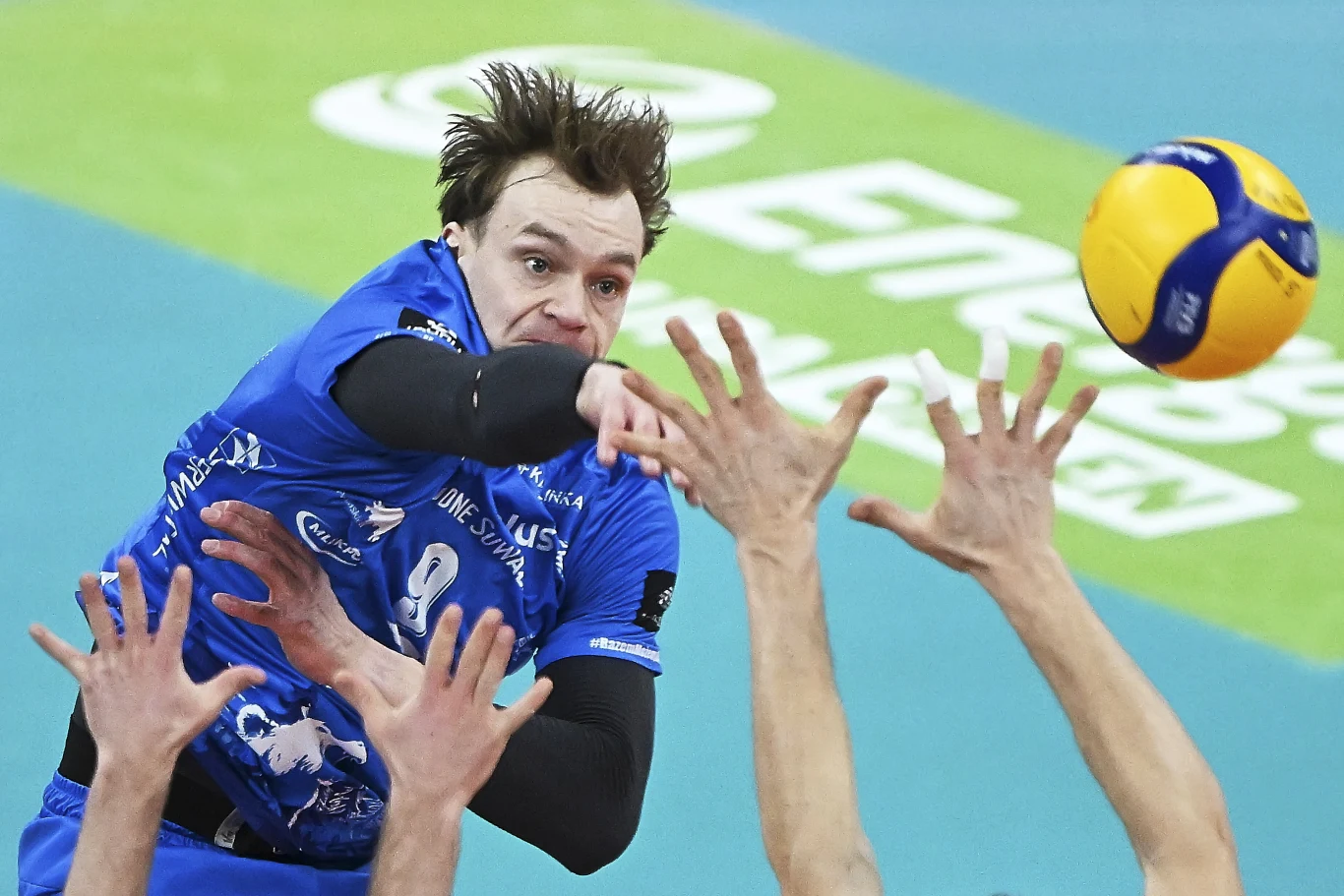 Bartosz Filipiak, atakujący Ślepska Malow Suwałki Zawodnik siatkarski w niebieskim stroju wyskakuje, wykonując atak nad siatką, podczas gdy po drugiej stronie widoczne są ręce próbujące go zablokować. W tle fragment boiska do siatkówki z widocznym logo sponsora.