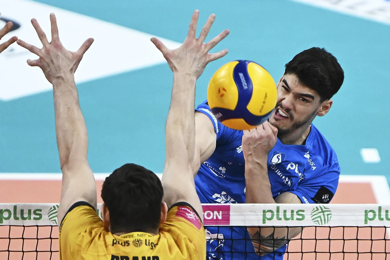 Henrique Honorato w meczu z Energa Treflem Gdańsk Zawodnik drużyny w niebieskim stroju wykonuje dynamiczny atak siatkarski nad siatką, podczas gdy przeciwnik w żółtej koszulce próbuje blokować piłkę unosząc wysoko ręce