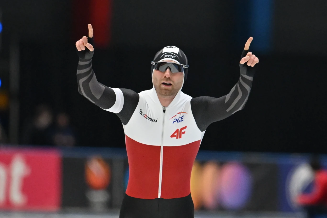 Łyżwiarz szybki w kombinezonie narodowym z podniesionymi rękami i uniesionymi palcami wskazującymi, wyrażający radość po zawodach na lodowisku.