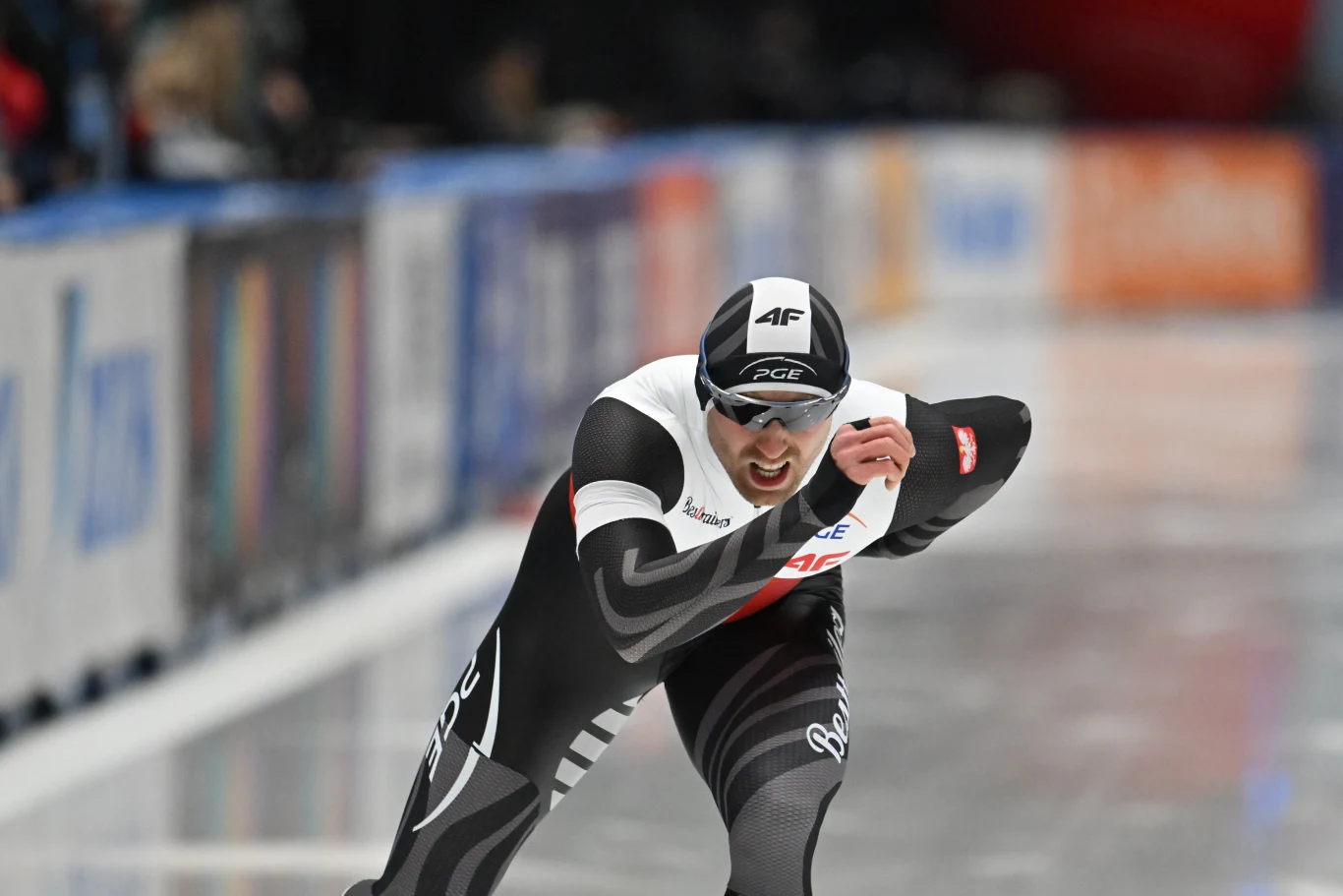 Łyżwiarz szybki w czarno-białym stroju startowym w dynamicznej pozie, ściga się po lodowej tafli, skupiony na rywalizacji, w tle rozmazane barwy trybun i reklam.