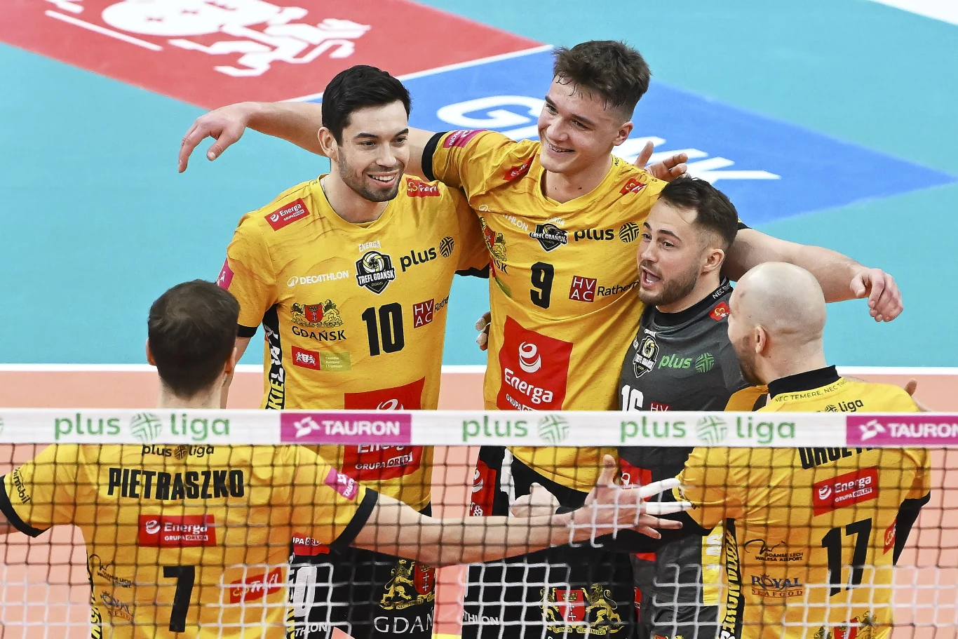 Siatkarze z Gdańska wyrwali zwycięstwo ze Ślepskiem Malow Suwałki Grupa siatkarzy w żółto-czarnych strojach PlusLigi świętuje wspólnie zdobyty punkt, stojąc po jednej stronie siatki na boisku do siatkówki. Zawodnicy uśmiechają się, obejmują i są w centrum uwagi, z trybunami w tle.