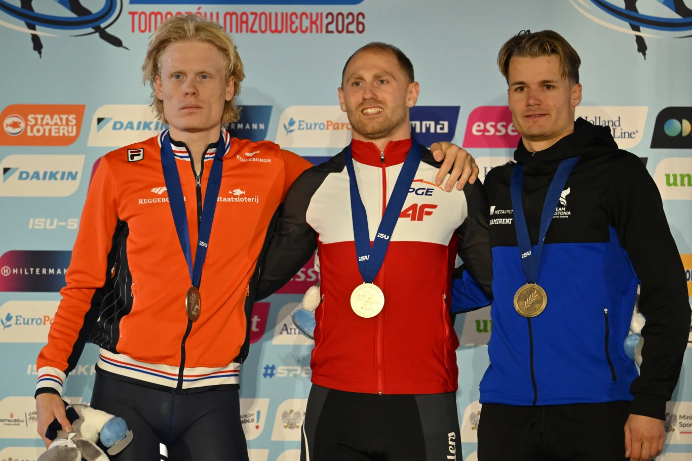 Trzech mężczyzn stojących na podium sportowym, każdemu zawieszony na szyi medal, tło z logo sponsorów oraz napisem Tomaszów Mazowiecki 2026, środkowy sportowiec trzyma rękę na ramieniu pozostałych.