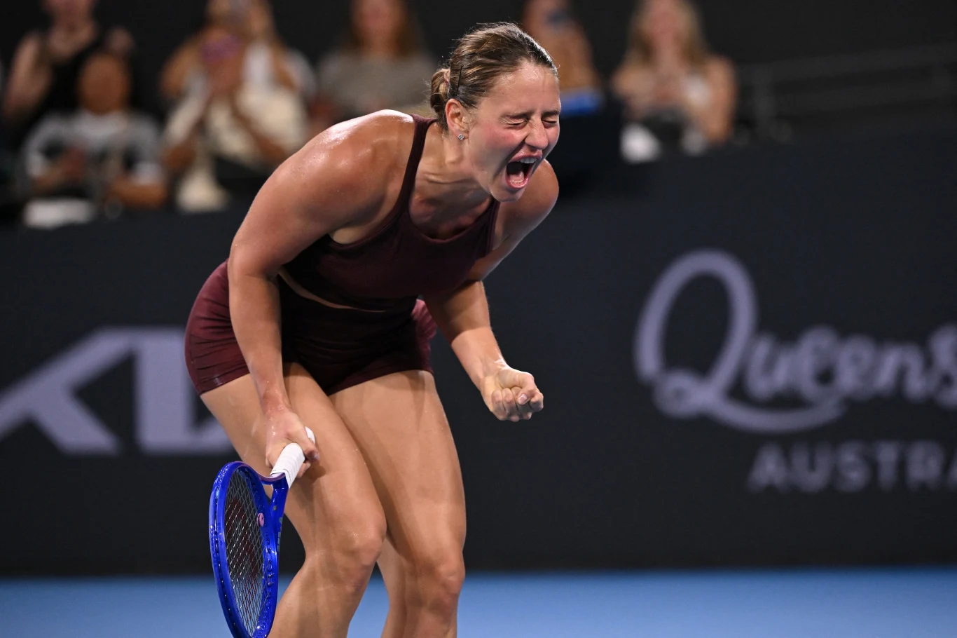 Marta Kostiuk, podopieczna polskiej trenerki Sandry Zaniewskiej, awansowała do finału WTA Brisbane Zawodniczka tenisowa w ciemnym stroju sportowym wykrzykuje z emocji, ściskając rakietę tenisową jedną ręką, na tle kortu i rozmytego napisu Queen’s Australia, wokół widoczna publiczność.