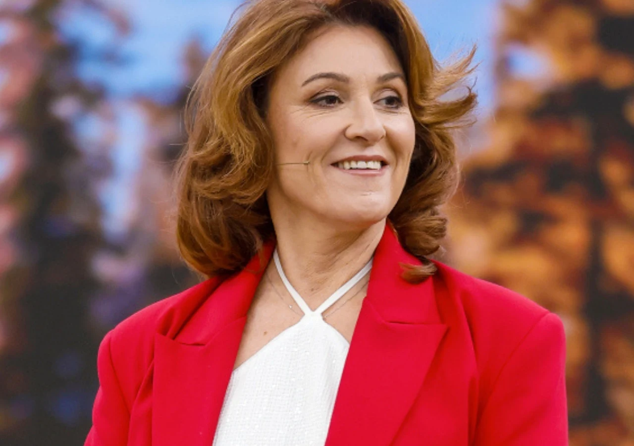 Agnieszka Hekiert Kobieta o brązowych falowanych włosach w średnim wieku, ubrana w biały top i elegancką czerwoną marynarkę, uśmiechnięta, stoi na tle rozmytego krajobrazu z drzewami o jesiennych barwach.