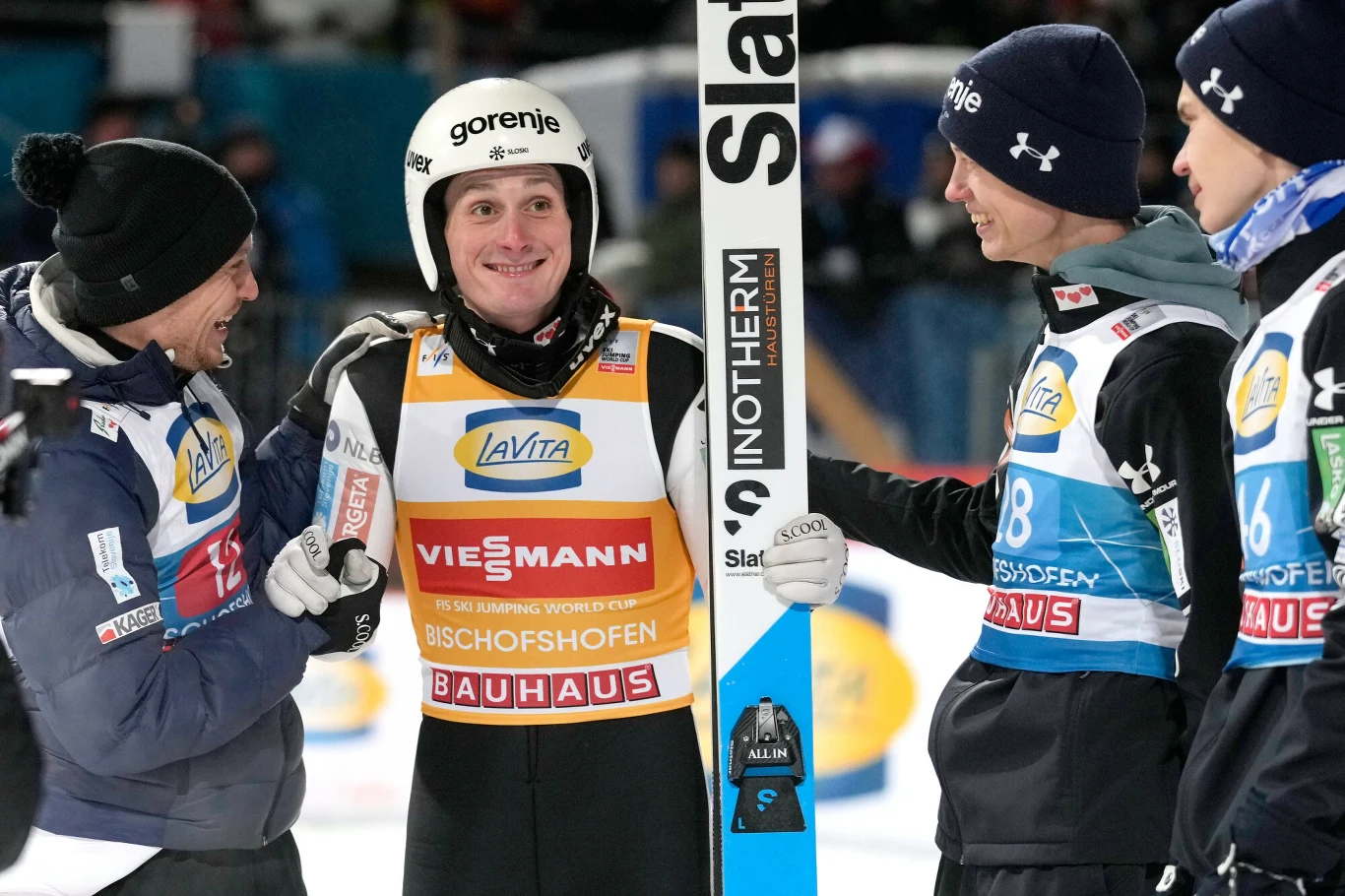 Skoczek narciarski w kasku i kombinezonie stojący wśród innych sportowców oraz trenera, trzyma narty, wszyscy uśmiechnięci celebrują sportowy sukces na tle zimowej areny.