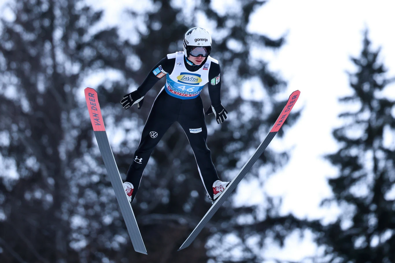 Zawodnik w kombinezonie sportowym wykonuje skok na nartach na tle rozmytych drzew, szeroko rozstawione narty w powietrzu, na głowie kask ochronny.