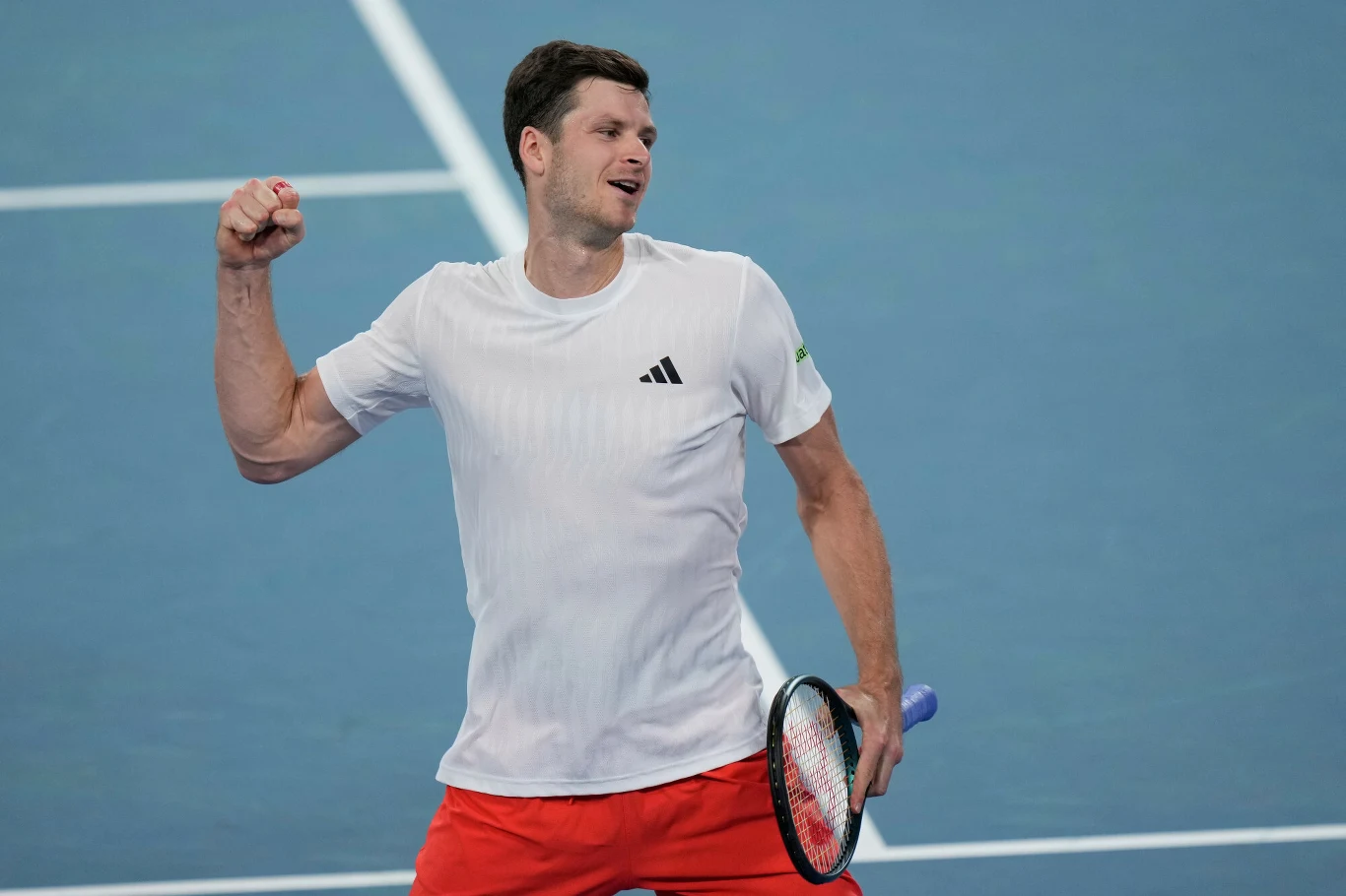 Tenisista w białej koszulce i czerwonych spodenkach świętuje zwycięski punkt na niebieskim korcie, trzymając rakietę do tenisa w prawej ręce.