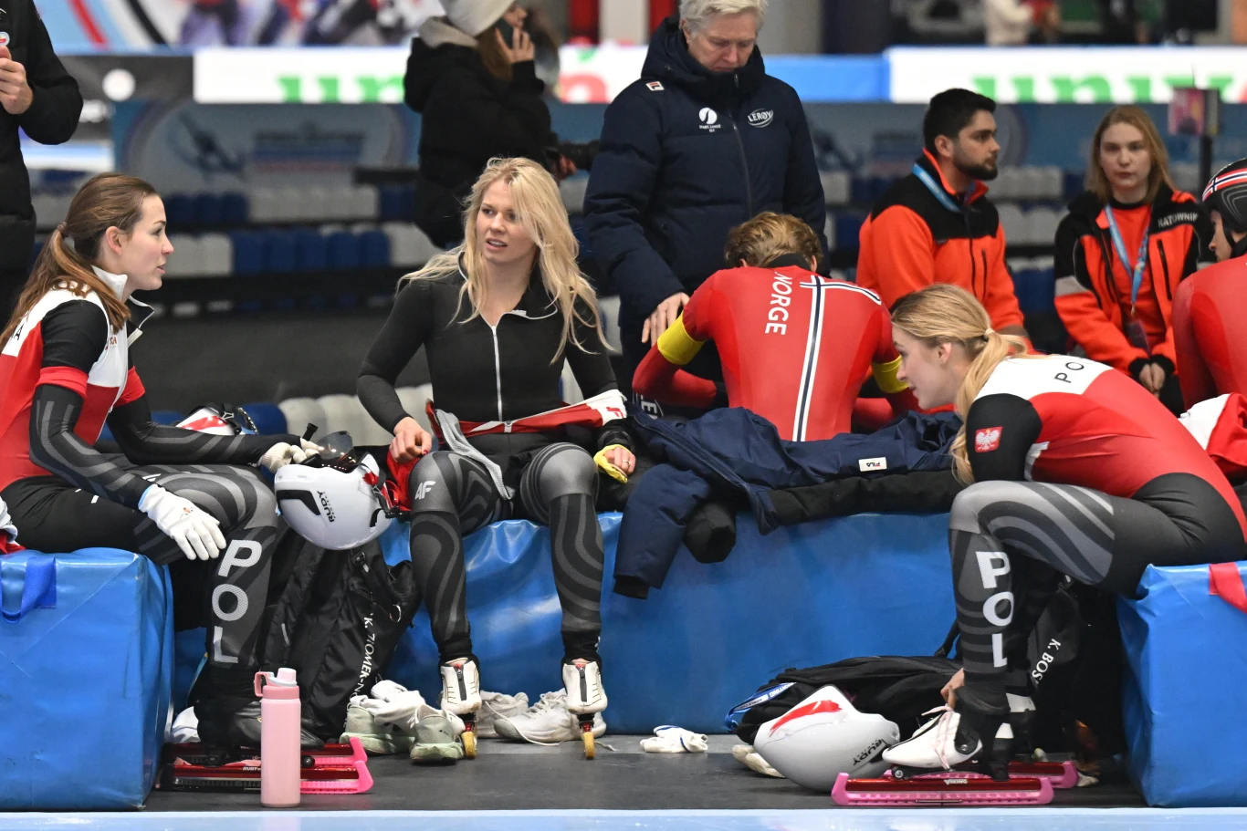 Zawodniczki w strojach do łyżwiarstwa szybkiego siedzą na niebieskich ławkach, wokół nich kaski i akcesoria sportowe, w tle trenerzy i obsługa oraz osoby w strojach sportowych.