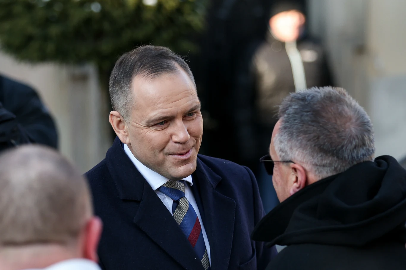 Karol Nawrocki na XVIII Patriotycznej Pielgrzymce Kibiców na Jasną Górę Mężczyzna w garniturze rozmawia z innym mężczyzną w okularach, w tle niewyraźna sylwetka oraz elementy architektury. Ubrania sugerują formalny lub oficjalny charakter sytuacji.