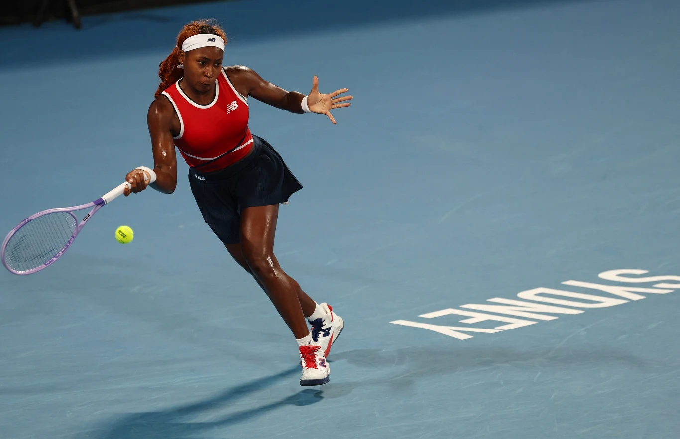 Tenisistka w czerwonym topie i granatowej spódnicy wykonuje forehand na kortach z niebieską nawierzchnią, piłka tenisowa zbliża się do niej, na podłodze widoczny biały napis SYDNEY.