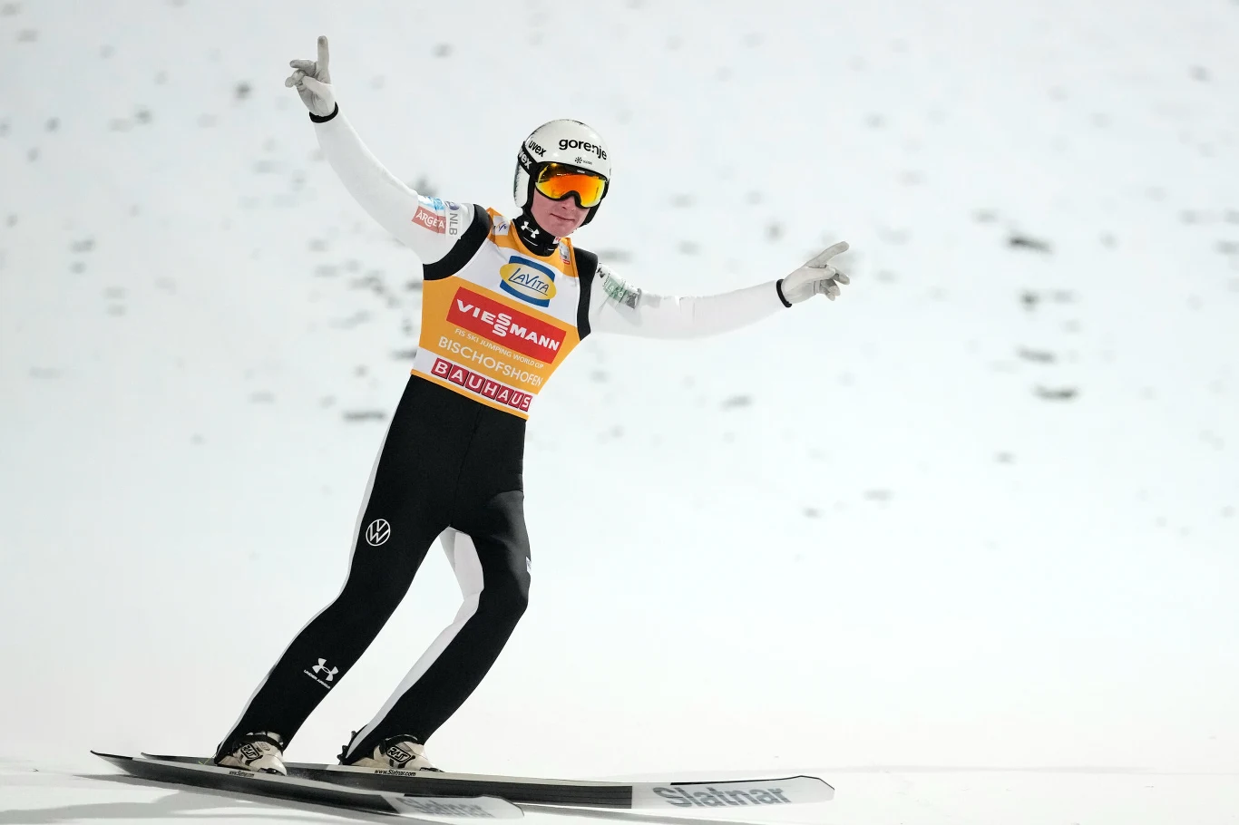 Skoczek narciarski ląduje na śnieżnej skoczni z rozpostartymi ramionami i uniesionymi dłońmi, ubrany w kombinezon sportowy oraz kask z goglami, tło stanowi jasna, zaśnieżona powierzchnia.