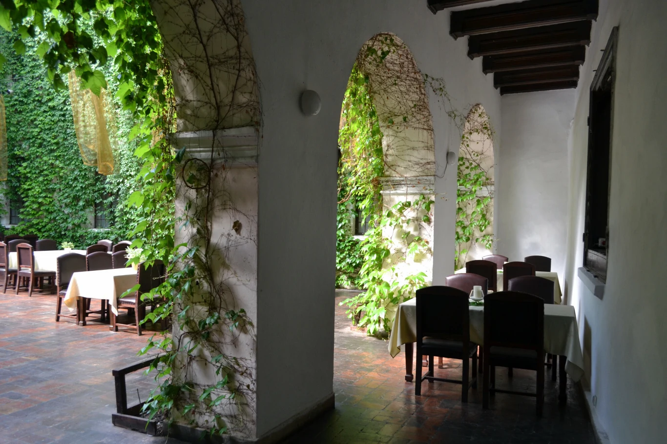 Korytarz z łukami prowadzący do części restauracyjnej na świeżym powietrzu, ściany porośnięte zielonymi pnączami, stoły nakryte białymi obrusami ustawione zarówno pod zadaszeniem, jak i w otwartym patio.