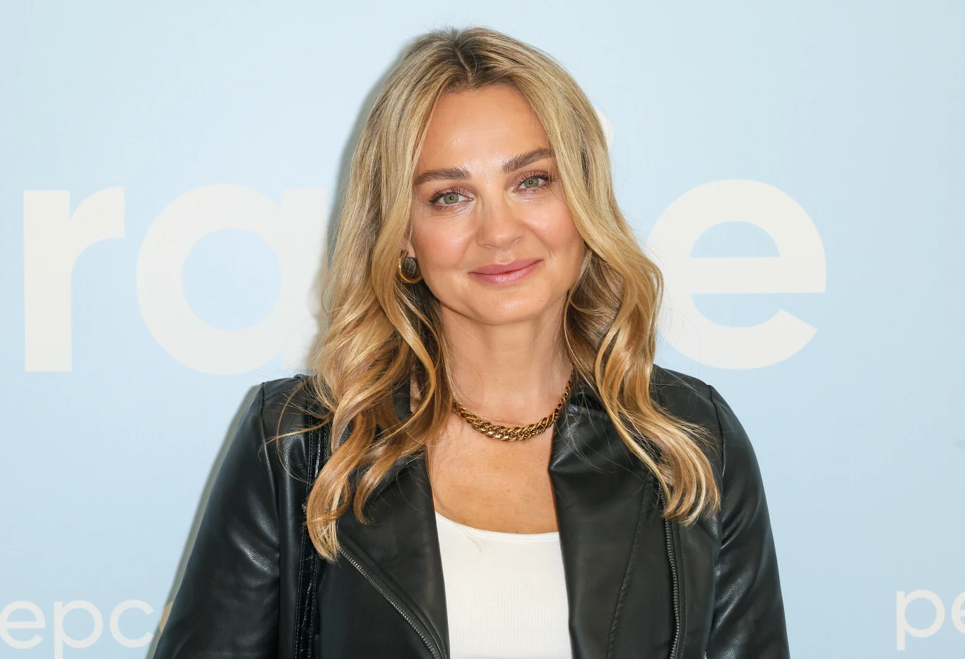 Małgorzata Socha odpoczywa na wakacjach Blondynka z długimi, falowanymi włosami, uśmiechnięta, ubrana w czarną skórzaną kurtkę oraz biały top, na jasnoniebieskim tle z białym napisem w tle.