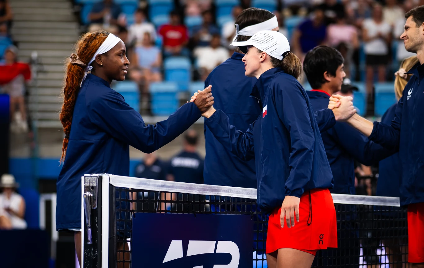 Dwie zawodniczki ubrane w sportowe stroje wymieniają uścisk dłoni przez siatkę na korcie tenisowym, w tle widoczni są inni sportowcy również witający się ze sobą oraz trybuny z widownią.