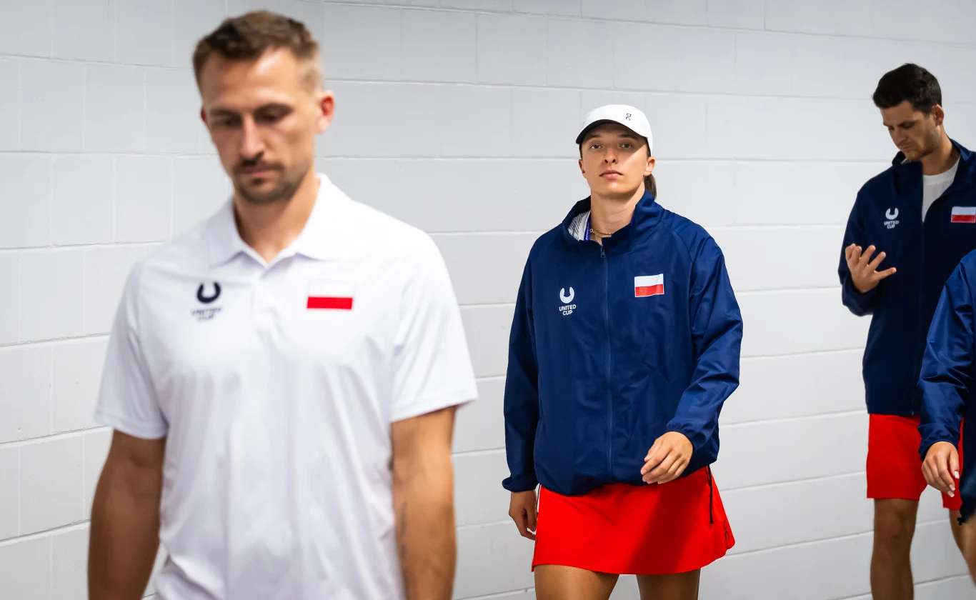 Iga Świątek, a obok niej: Jan Zieliński (z lewej) i Hubert Hurkacz (z prawej) Troje sportowców ubranych w stroje z polskimi flagami idzie korytarzem. Kobieta na środku ma granatową kurtkę, czerwoną spódnicę i białą czapkę, mężczyzna z przodu jest w białej koszulce, a mężczyzna z tyłu w granatowej kurtce i czerwonych spodenkach. ...