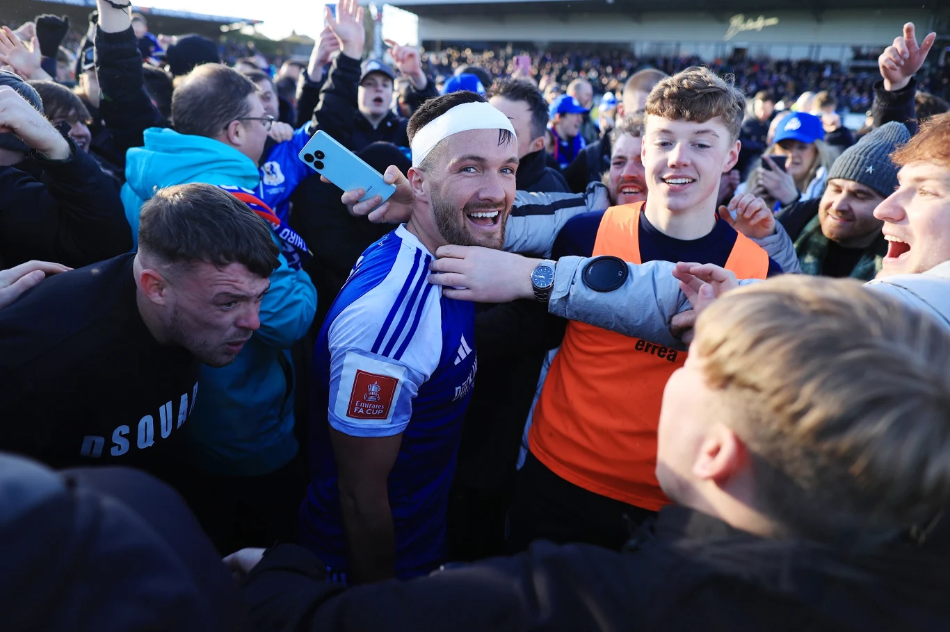 Tłum kibiców otacza zawodnika w sportowym stroju z opaską na głowie, który uśmiecha się szeroko, unosząc ręce w geście zwycięstwa, w tle widać pełne trybuny stadionu.