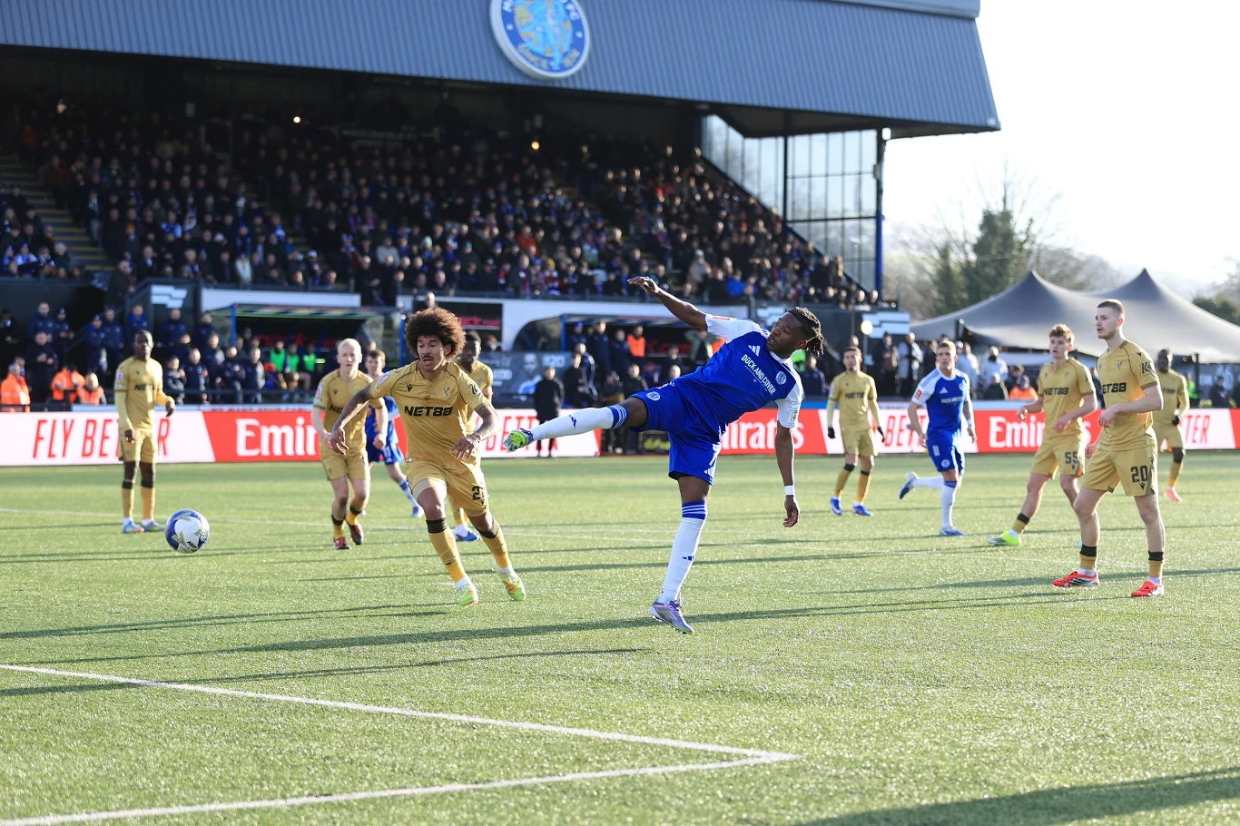 Piłkarz w niebieskim stroju wykonuje dynamiczny strzał na bramkę podczas meczu, wokół niego kilku zawodników w żółtych koszulkach, w tle zapełnione trybuny oraz stadion.