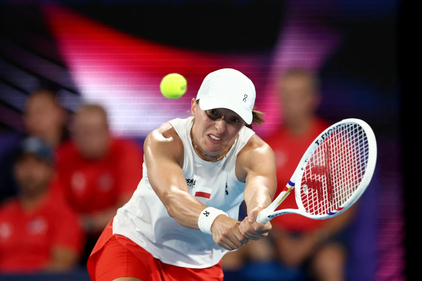 Tenisistka w białej czapce i stroju sportowym odbija piłkę rakietą podczas intensywnego meczu. W tle rozmazane sylwetki osób oraz kontrastujący, kolorowy element.