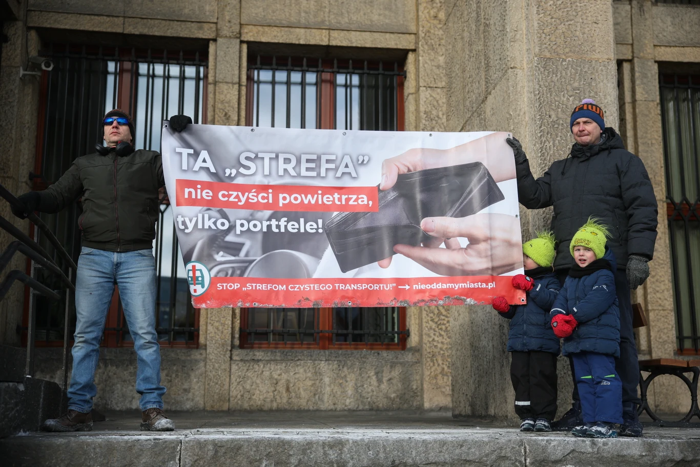 Protestujący trzymają transparent z napisem krytykującym strefę czystego transportu, sugerującym, że dotyczy ona bardziej pieniędzy niż jakości powietrza. Obok dorosłych stoją dzieci ubrane w zimowe stroje. Scena rozgrywa się na tle kamiennego budynku.