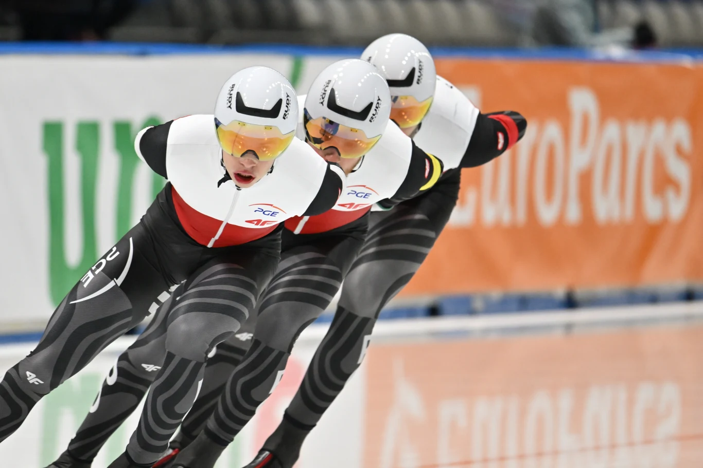 Trzech łyżwiarzy szybkich w identycznych kombinezonach ściga się na torze lodowym, pochylając się dynamicznie podczas jazdy, w tle widać niewyraźny baner reklamowy.