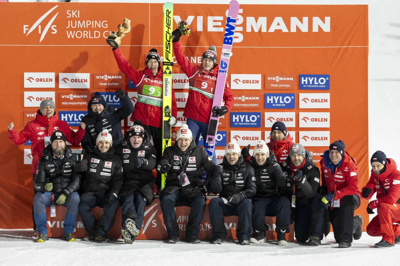 Polscy skoczkowie zakończyli konkurs duetów na trzecim miejscu Grupa skoczków narciarskich wraz z zespołem technicznym pozuje na podium na tle pomarańczowej ścianki sponsorskiej, dwóch zawodników stoi z nartami uniesionymi w geście radości, pozostali członkowie zespołu siedzą i klęczą uśmiechając się do kamery.