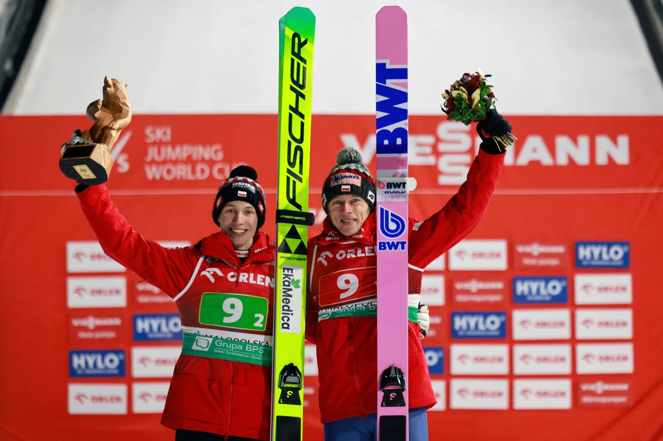 Dwóch skoczków narciarskich w czerwonych kurtkach, uśmiechniętych, trzymających narty oraz trofeum i bukiet kwiatów, stojących na tle czerwonej ścianki sponsorskiej podczas zawodów Pucharu Świata.