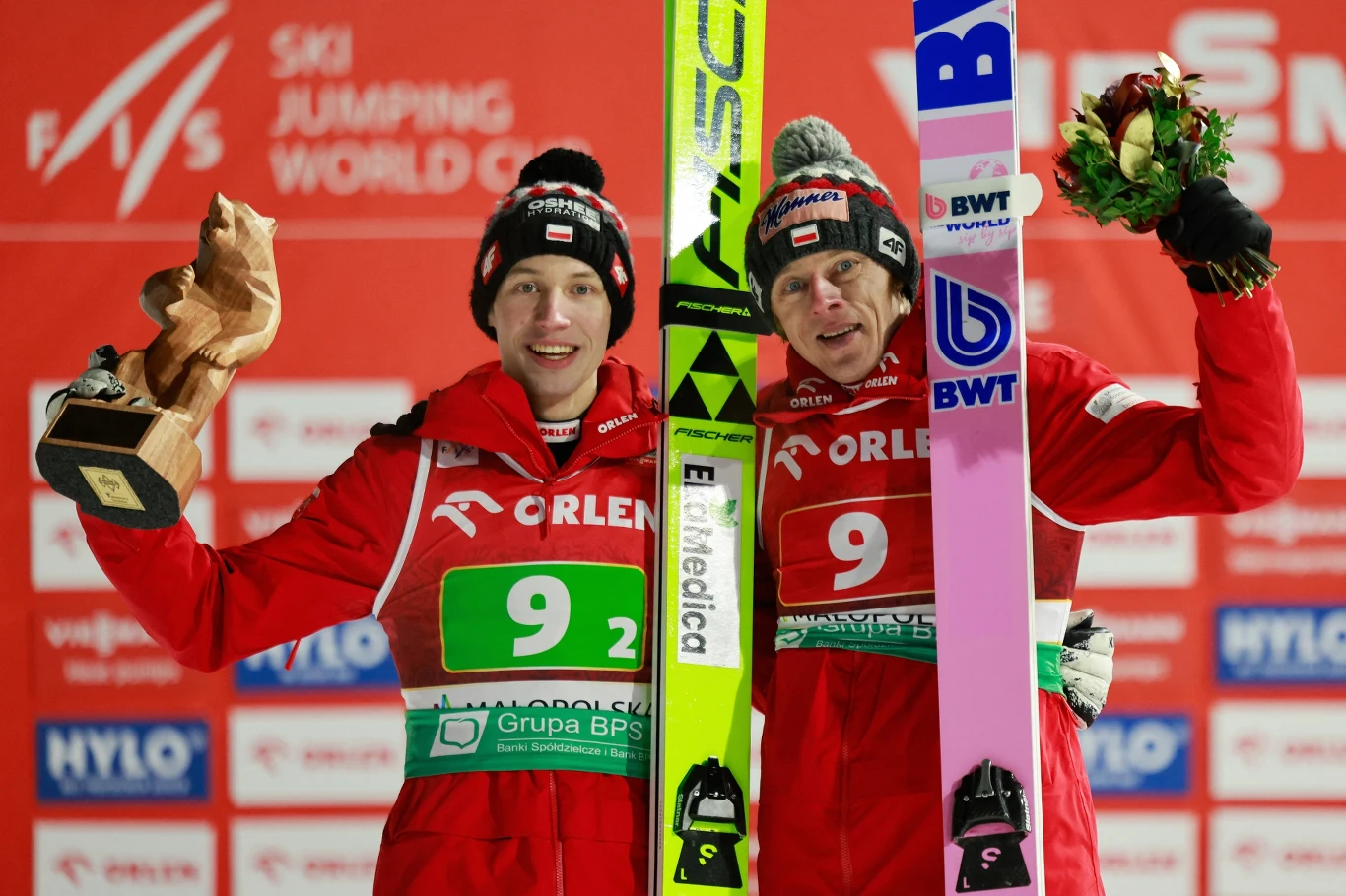 Polscy skoczkowie zajęli trzecie miejsce w konkursie duetów Dwóch polskich skoczków narciarskich stoi na podium z nartami, ubrani w czerwone kombinezony z logo sponsorów, jeden z nich trzyma drewnianą statuetkę, drugi bukiet kwiatów, w tle ścianka sponsora i logo zawodów Pucharu Świata.