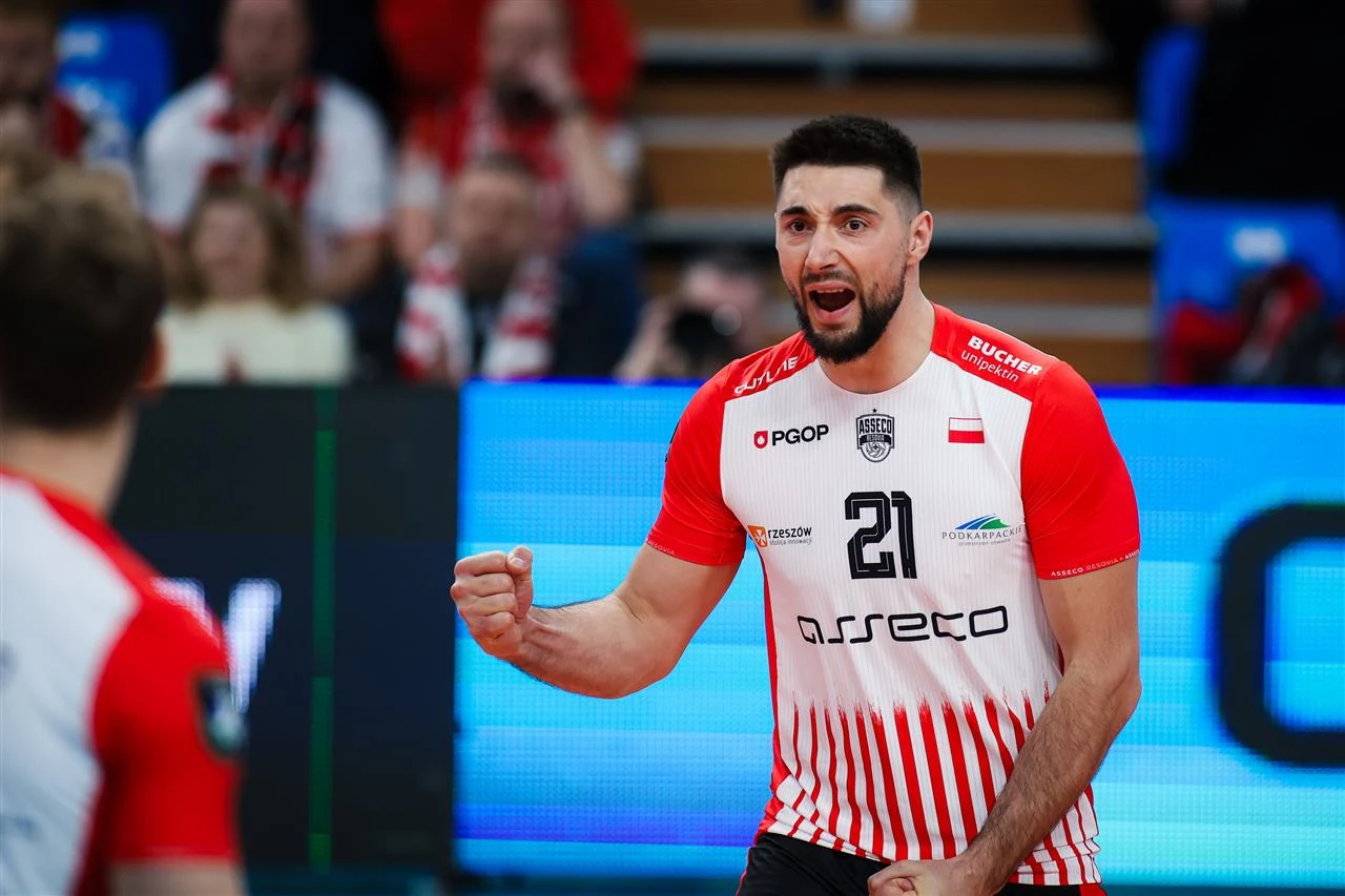 Zawodnik drużyny siatkarskiej w czerwono-białym stroju świętuje zdobycie punktu, widoczny na pierwszym planie z zaciśniętą pięścią i wyraźnym wyrazem radości na twarzy, w tle rozmyte sylwetki kibiców i fragment hali sportowej.