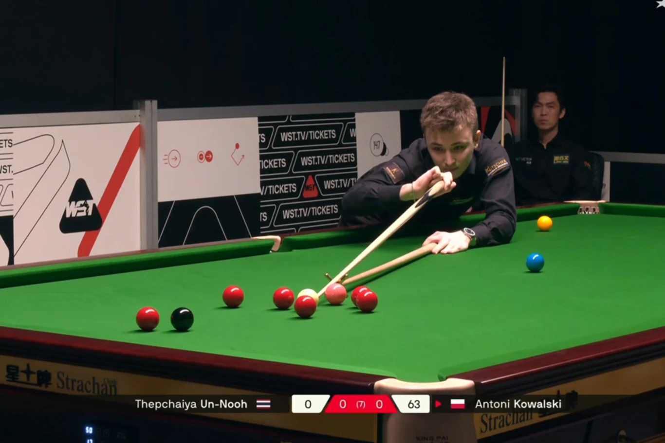 Tu pomylił się Antoni Kowalski. I kosztowało go to całego frejma Zawodnik podczas gry w snookera pochylony nad stołem wykonuje uderzenie kijem bilardowym. Na stole widoczne są czerwone, czarna, zielona i niebieska bila, a obok niego siedzi sędzia. W tle tablice wyników wskazują na trwający mecz pomiędzy Thepchaiya U...