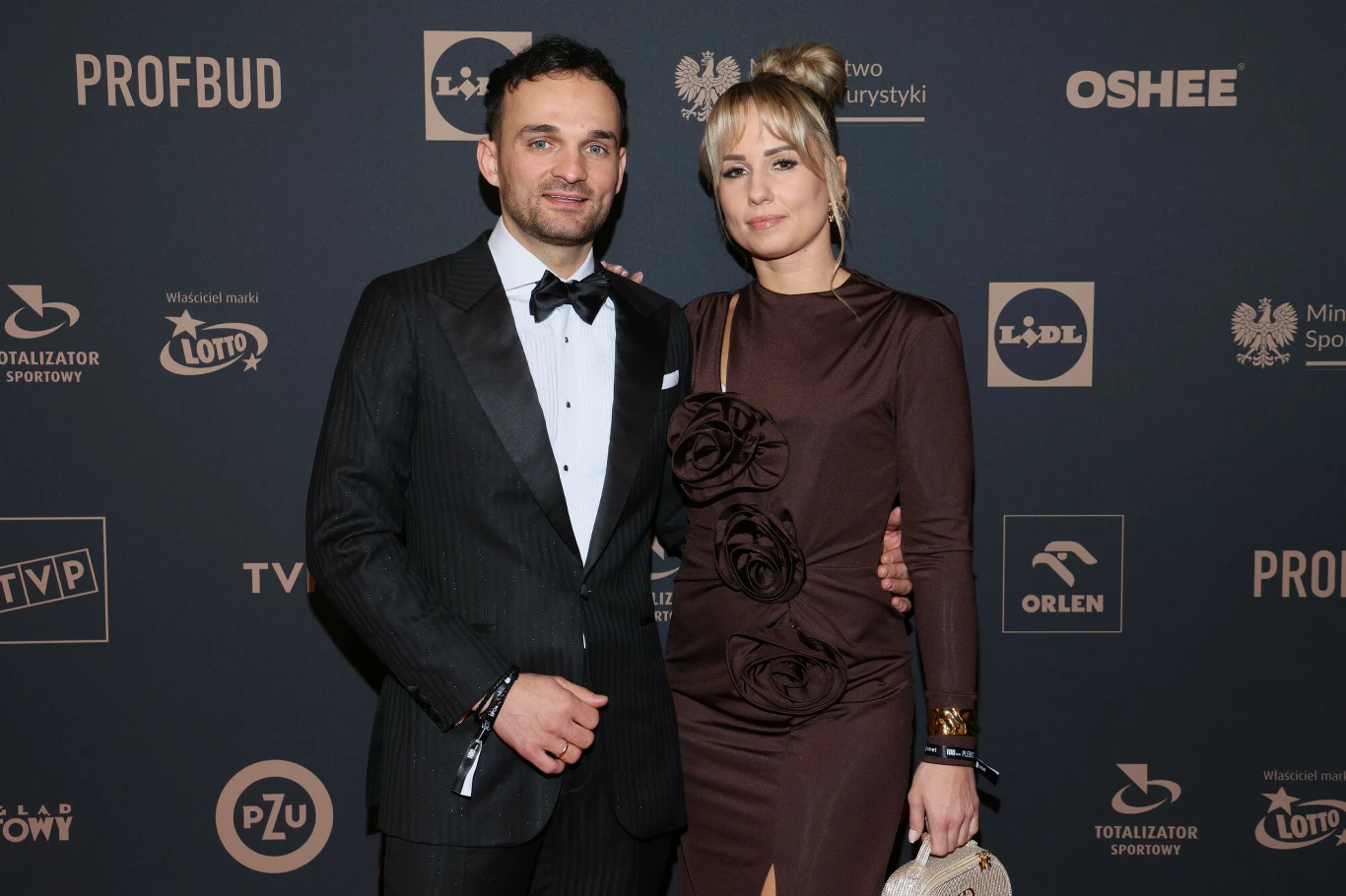 Elegancko ubrana para na tle ścianki sponsorskiej podczas oficjalnego wydarzenia. Mężczyzna w czarnym garniturze i muszce, kobieta w ciemnej sukni z kwiatowymi aplikacjami, trzyma srebrną torebkę. Oboje stoją blisko siebie i patrzą w stronę aparatu.